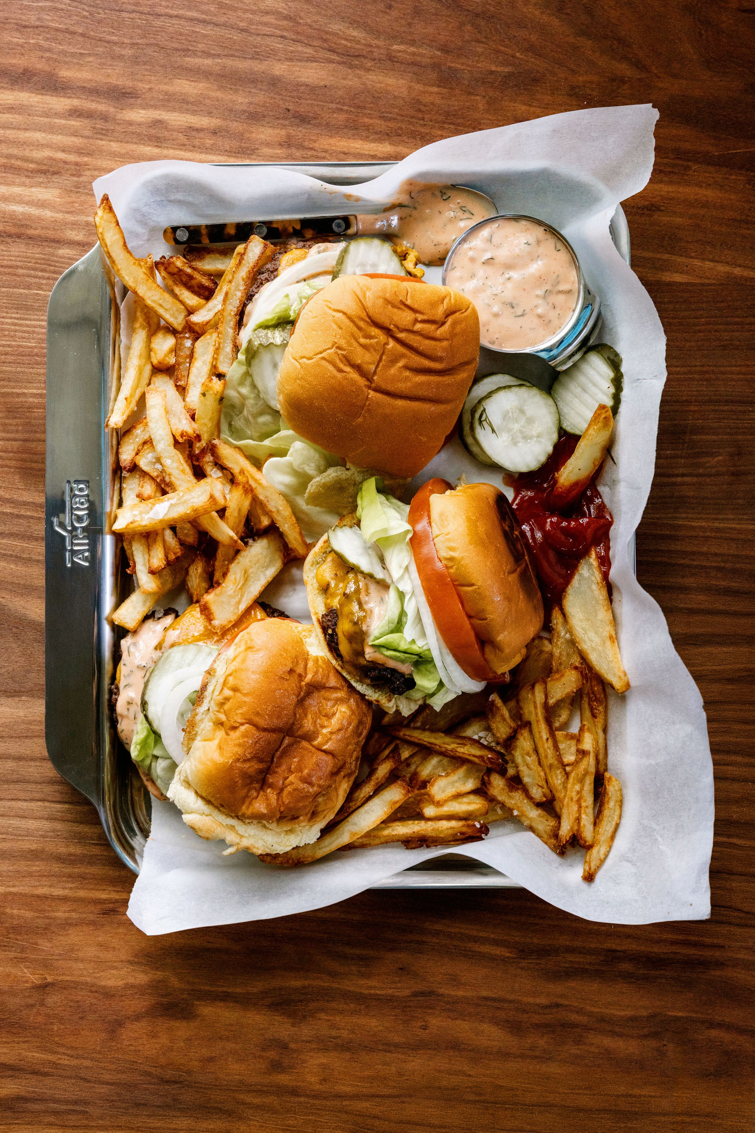 Two burgers with lettuce on brioche buns served with crispy fries and dipping sauce on an All-Clad tray.