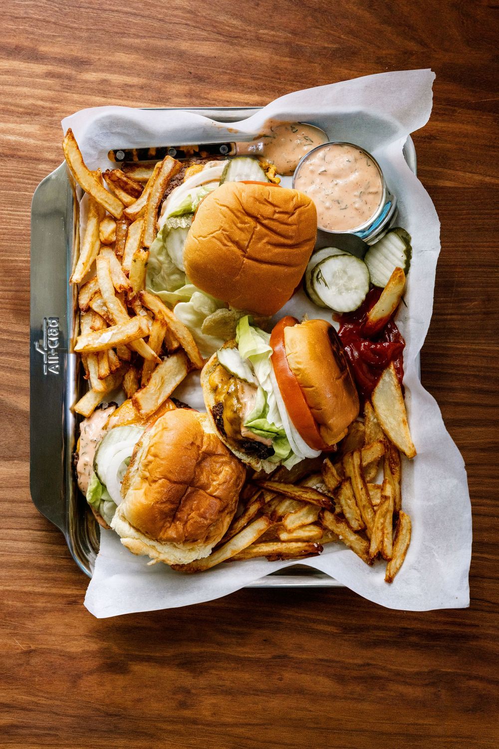 Two burgers with lettuce on brioche buns served with crispy fries and dipping sauce on an All-Clad tray.