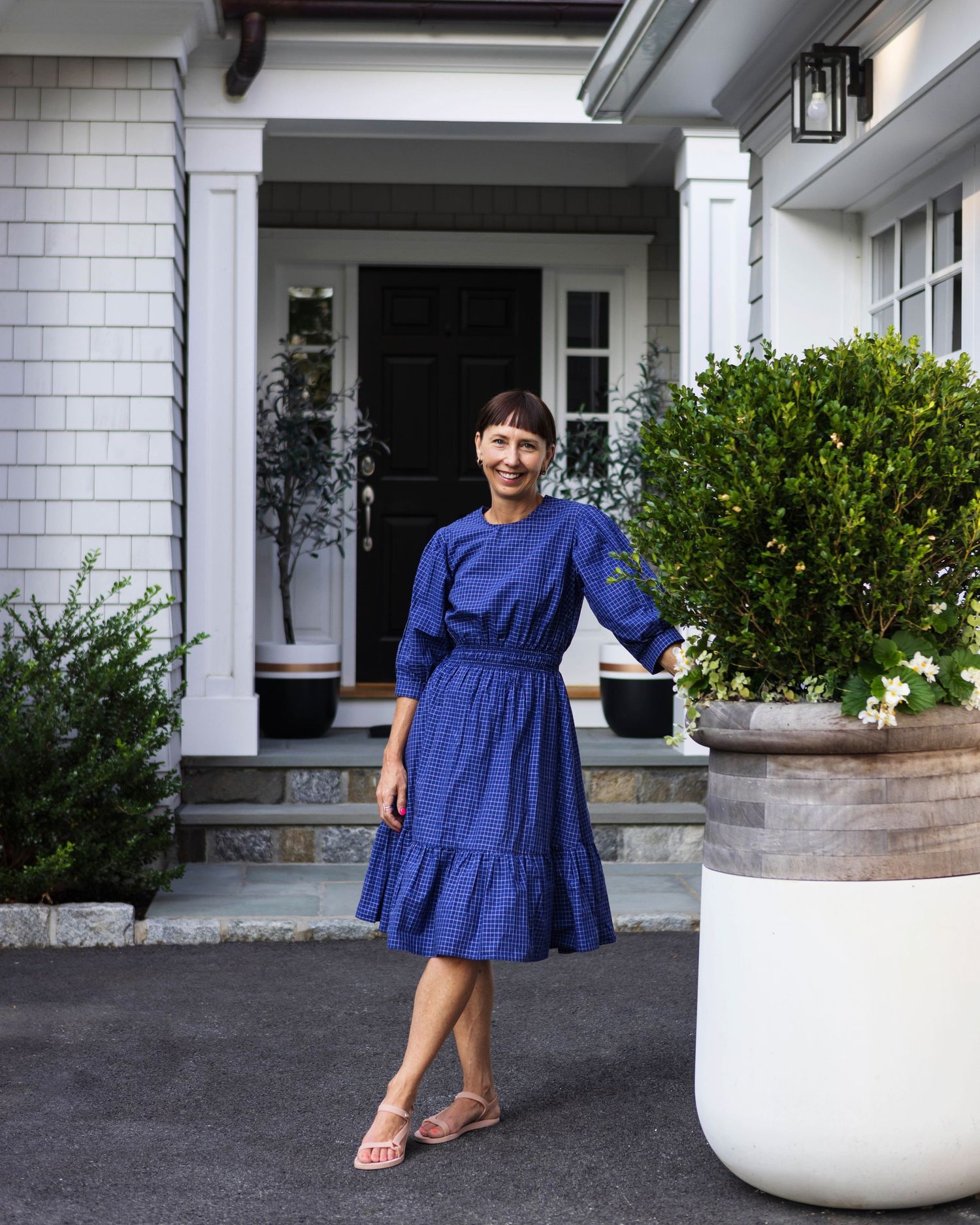 Woman in blue midi dress standing outside a charming gray shingle home with black door and lush greenery.