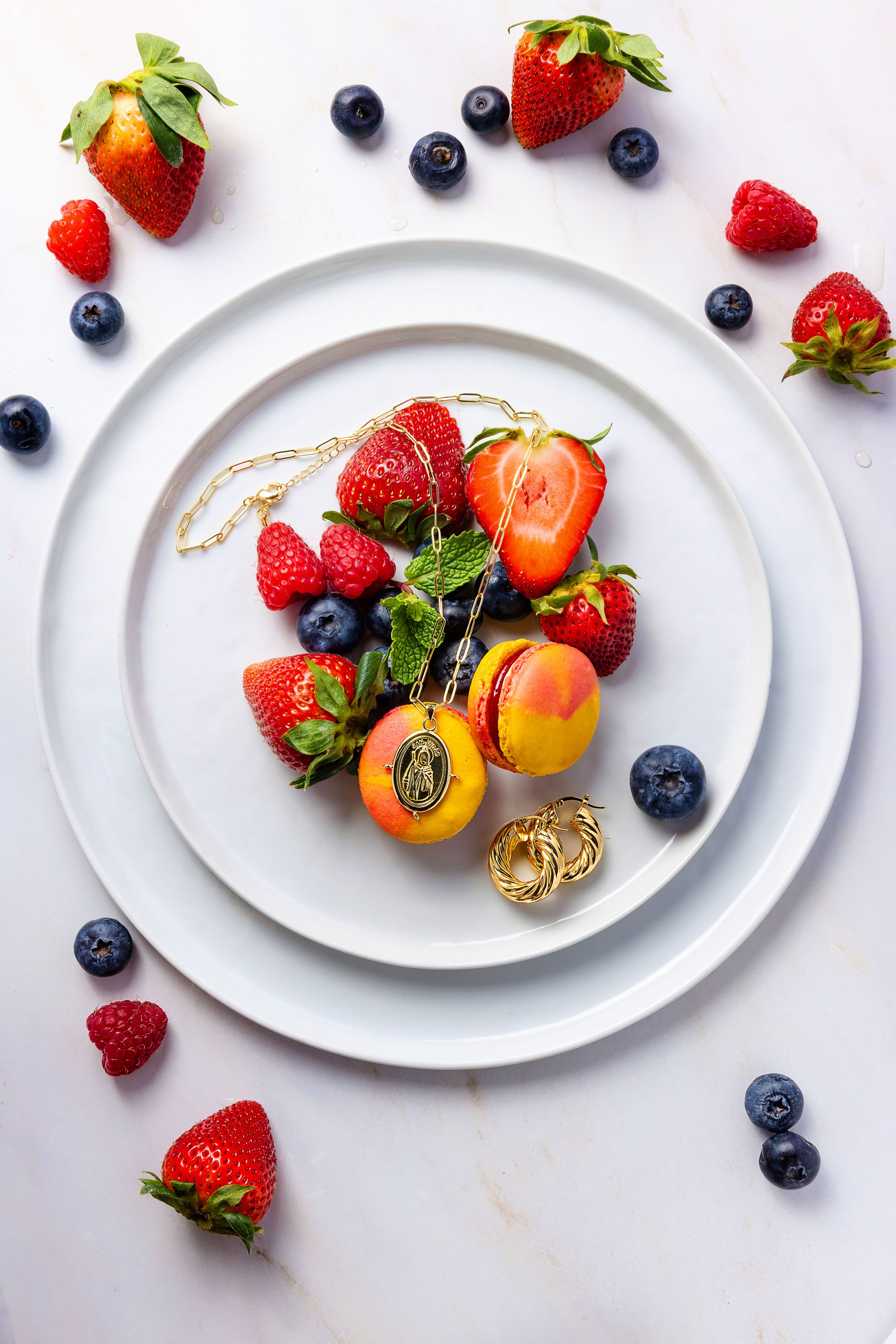 Fresh strawberries, raspberries, blueberries, and apricots arranged with gold jewelry on white plates.