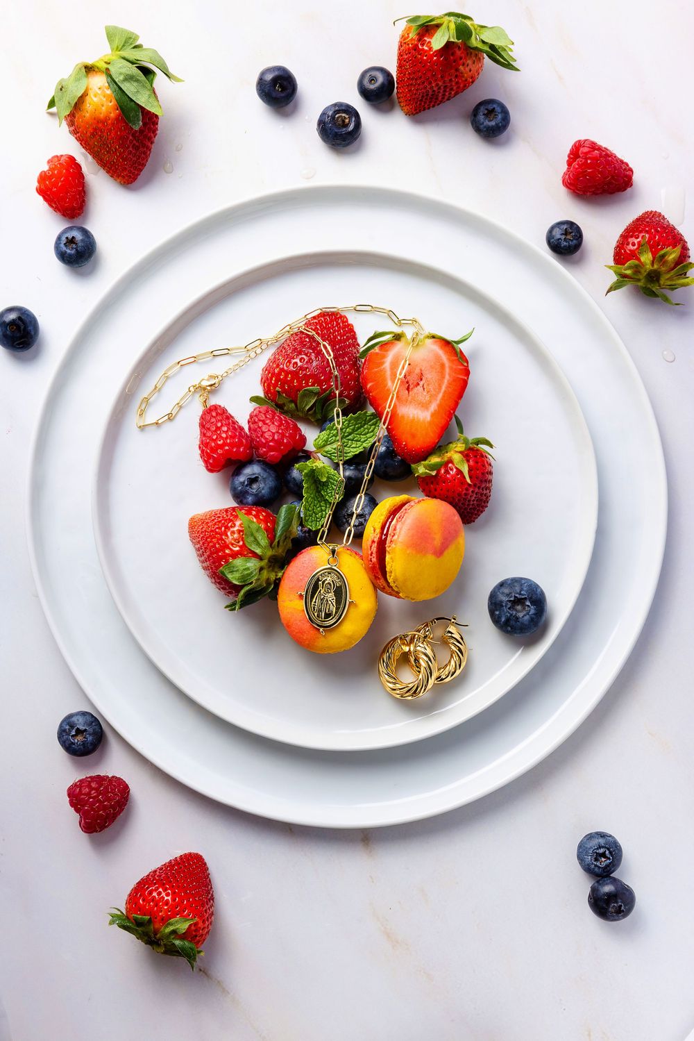Fresh strawberries, raspberries, blueberries, and apricots arranged with gold jewelry on white plates.