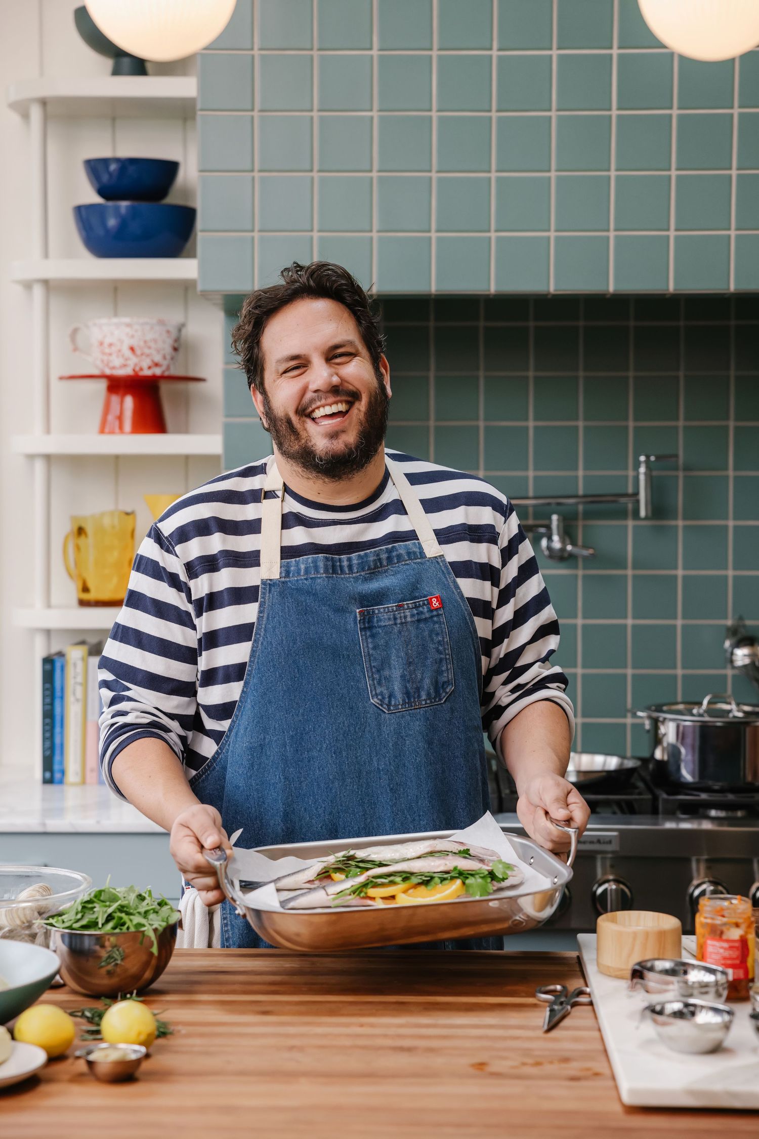 Smiling man in denim apron holds baking dish with vegetables in a modern kitchen with teal tiles.