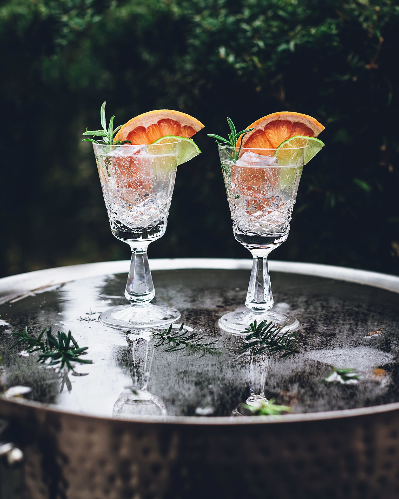 Two crystal goblets with crushed ice and citrus garnish sitting on a wet silver tray outdoors.