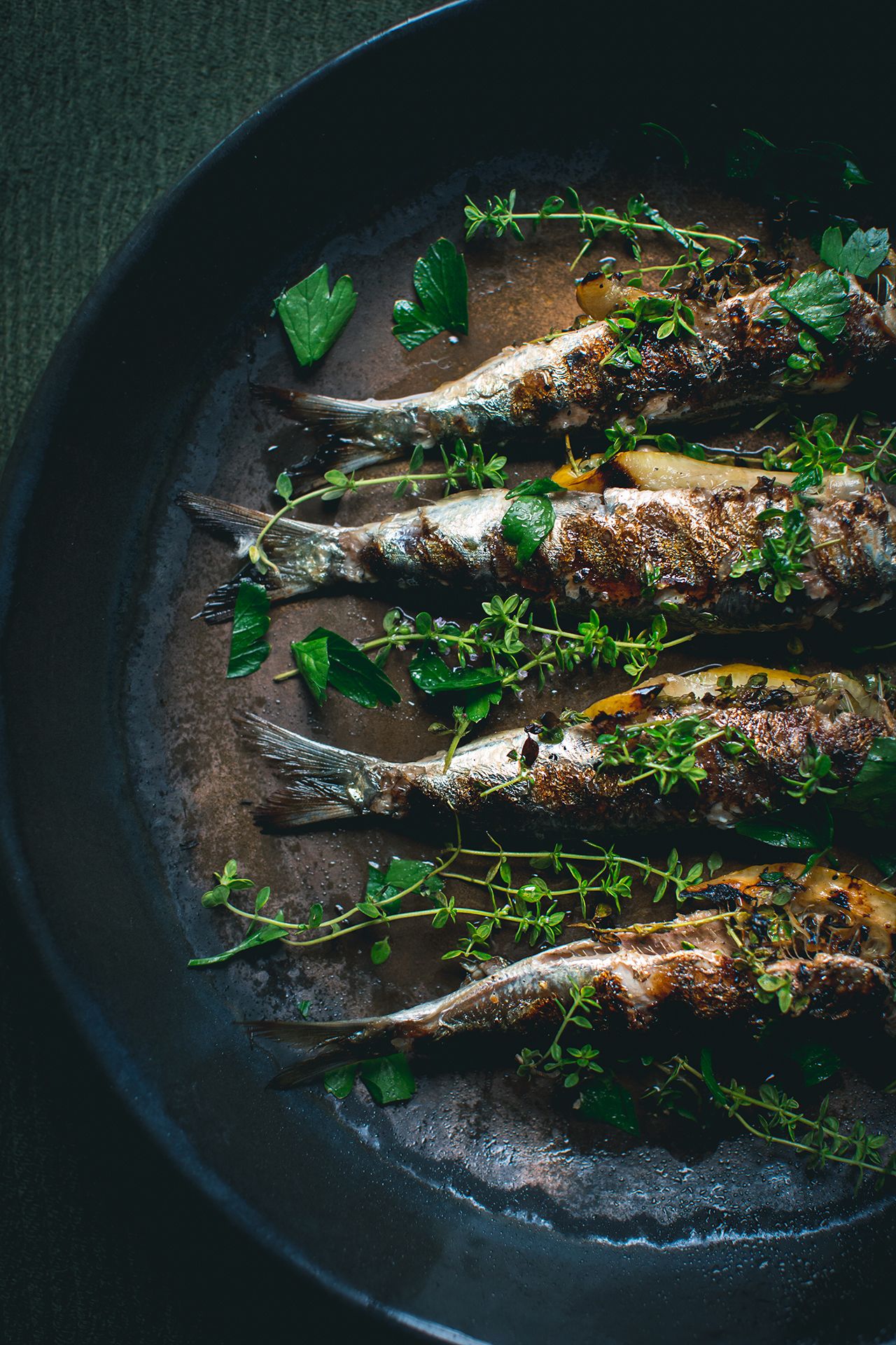 Four herb-topped grilled sardines resting in savory juices inside a dark cast iron skillet.