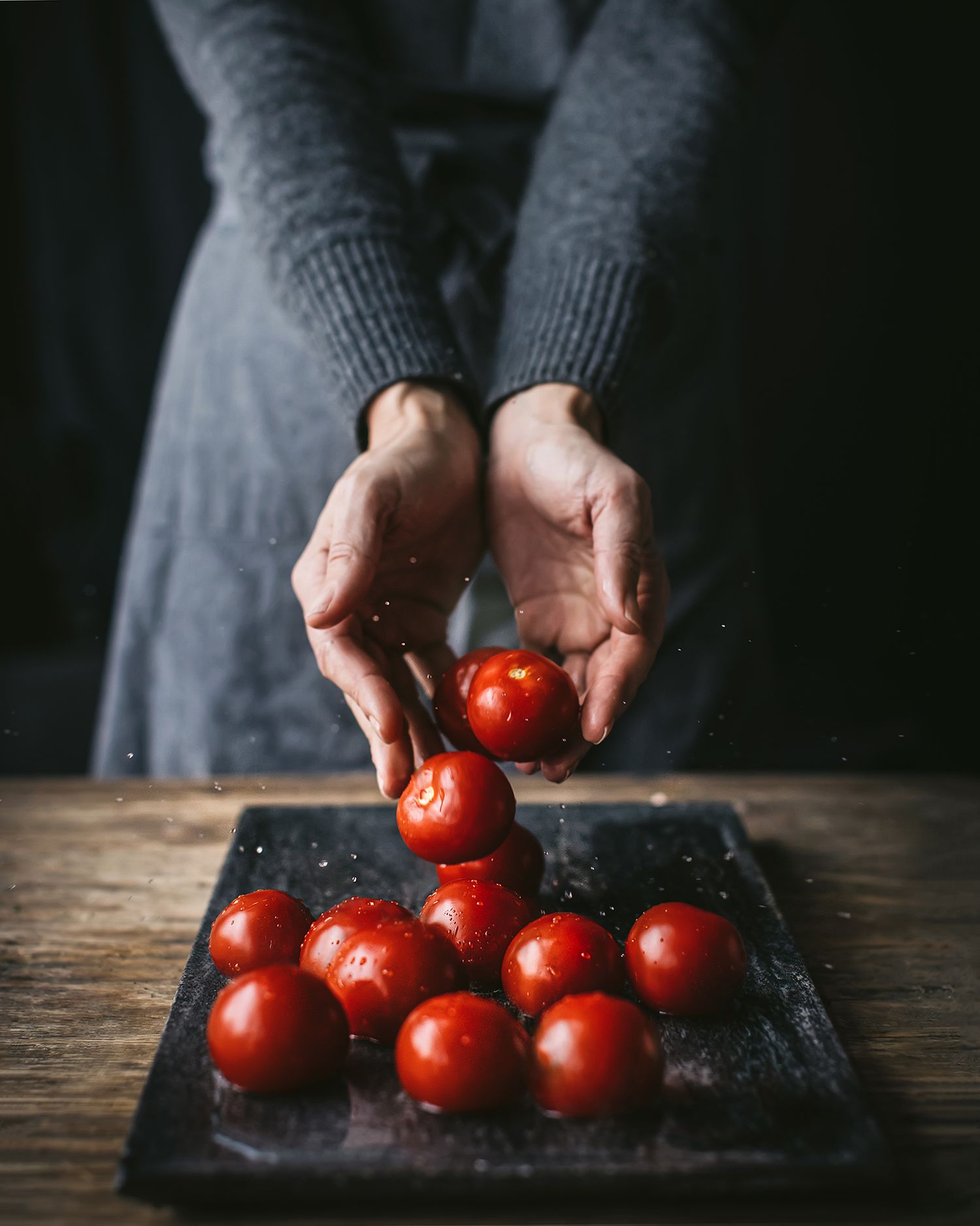 Person in gray sweater holding fresh red cherry tomatoes over a dark slate board on a rustic wooden table.