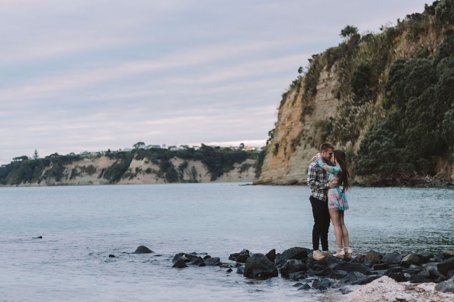 Couple shares an intimate moment on rocky beach with dramatic coastal cliffs in background.
