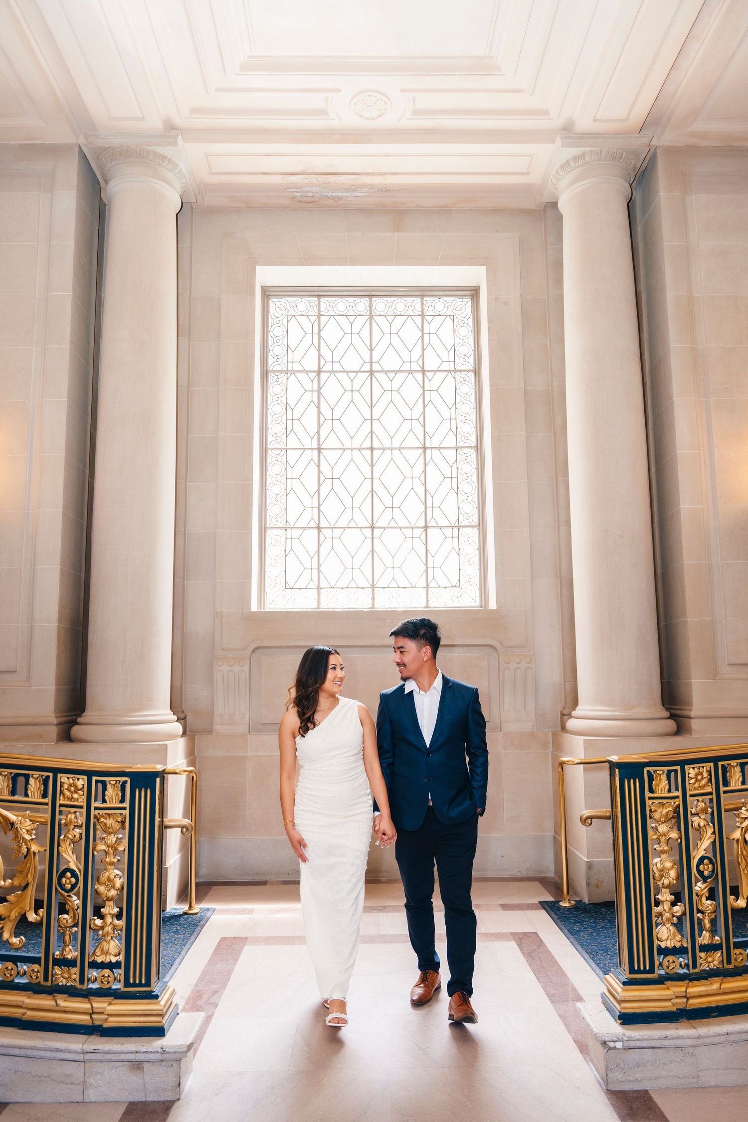 Couple in formal attire stands between elegant columns and gold railings in historic building.
