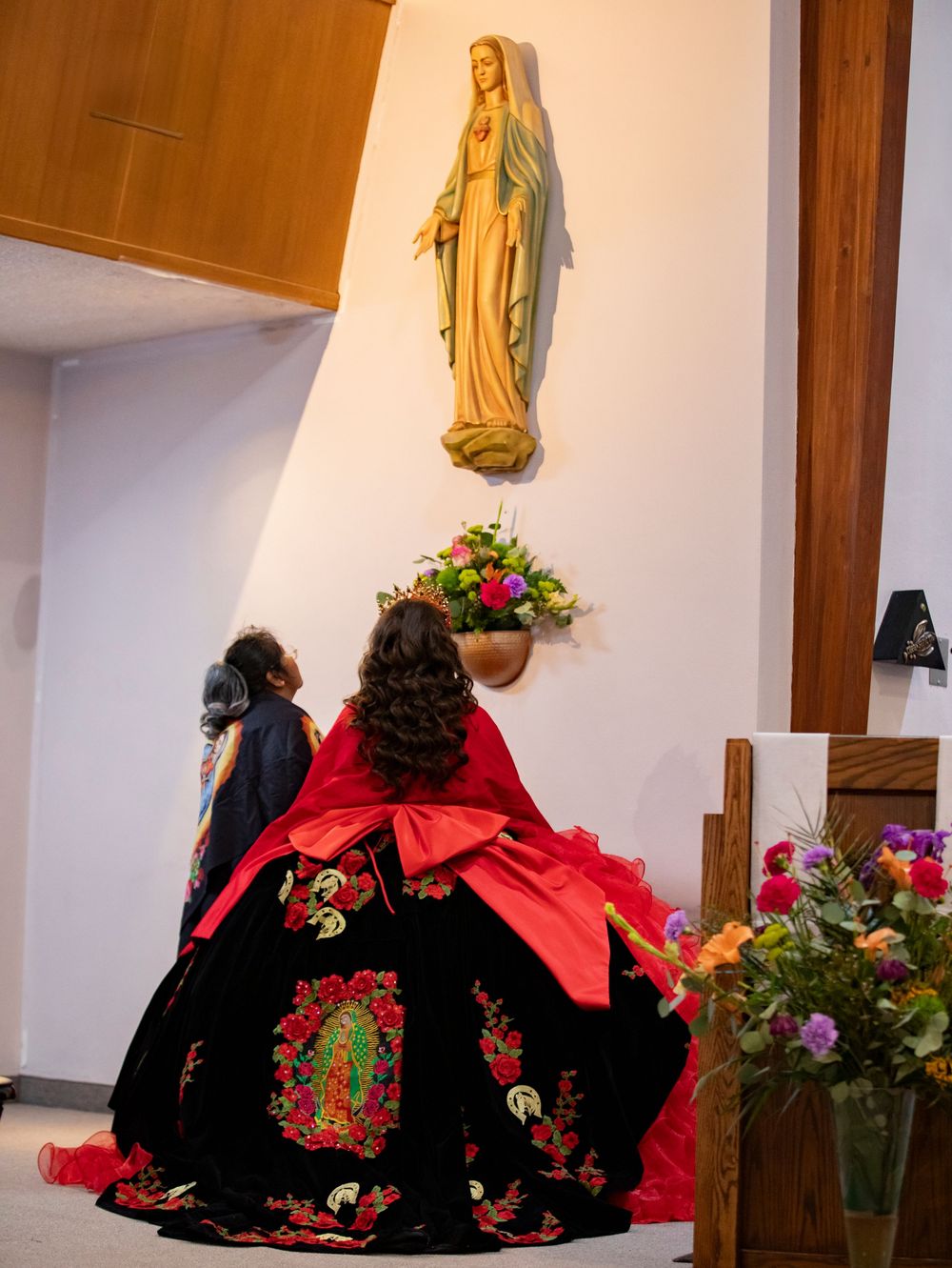 Religious statues adorn a church altar with red and black traditional Mexican dress and floral decorations.