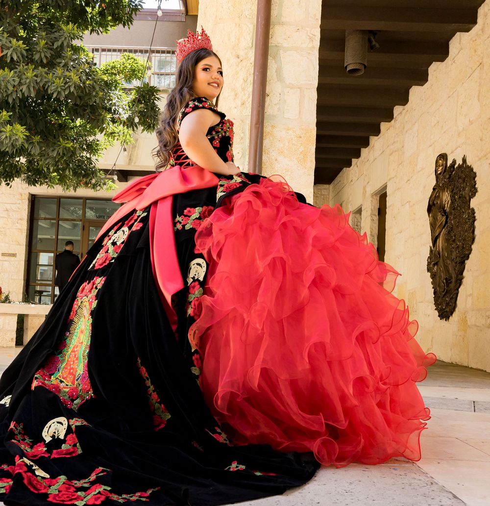 A person in a black and red quinceañera dress poses against a beige stone wall in an outdoor courtyard setting.