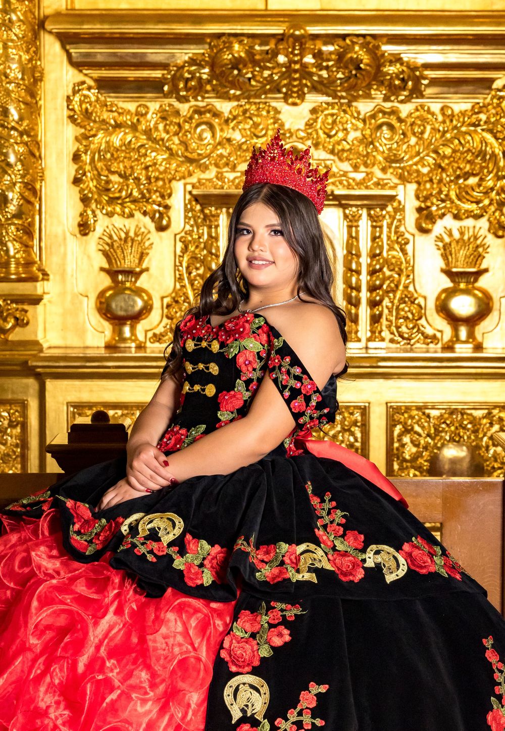 Ornate Mexican quinceañera dress in black and red with floral embroidery against an elegant gold backdrop.