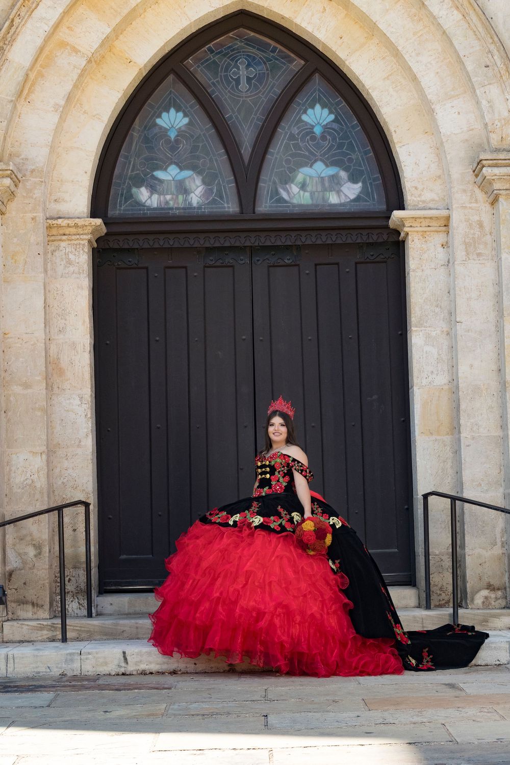 A dramatic red quinceañera dress displayed against ornate stone archways with black doors.