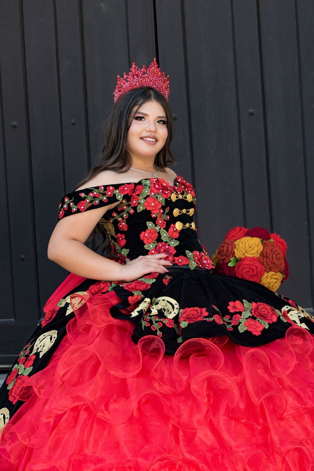 A vibrant red quinceañera dress with black embroidery and ruffled layers photographed against a dark background.