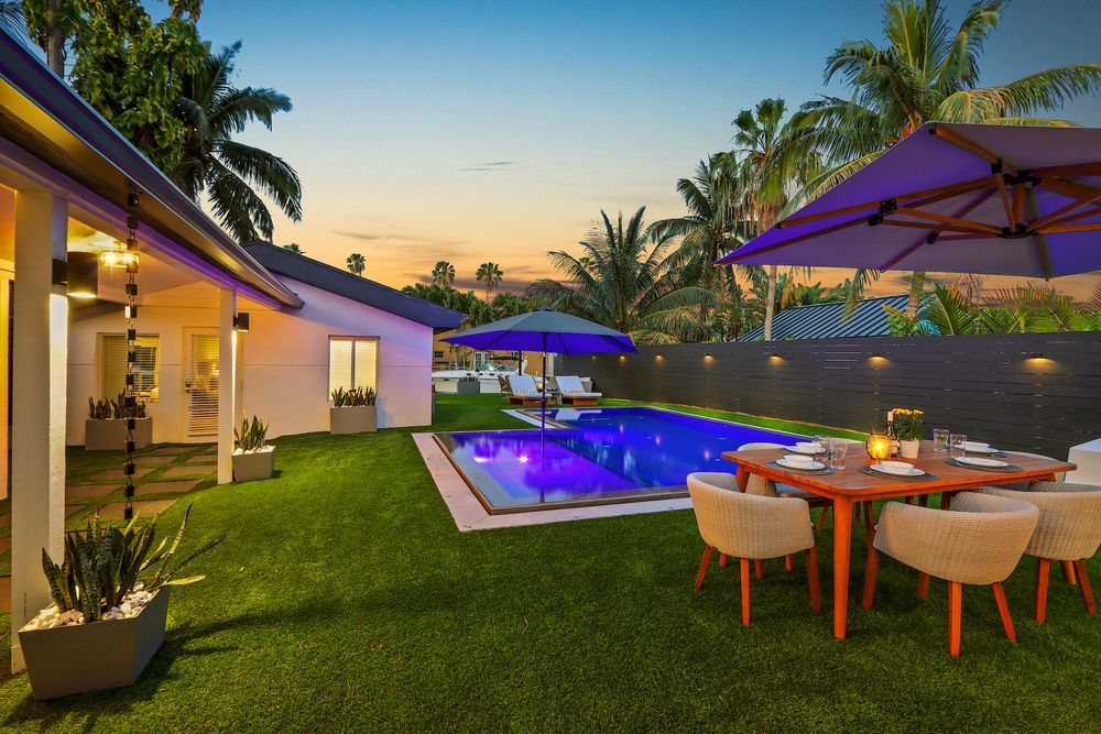 Modern backyard oasis with illuminated pool, palm trees, and outdoor dining area at sunset.