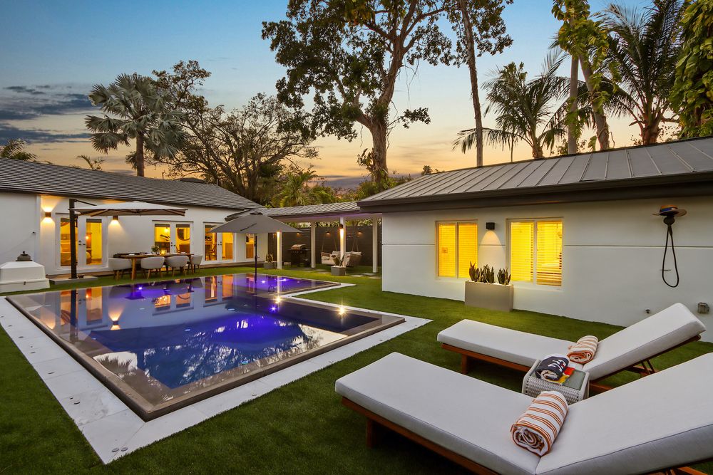 Modern backyard with illuminated swimming pool, lounge chairs and palm trees at sunset against a white luxury residence.