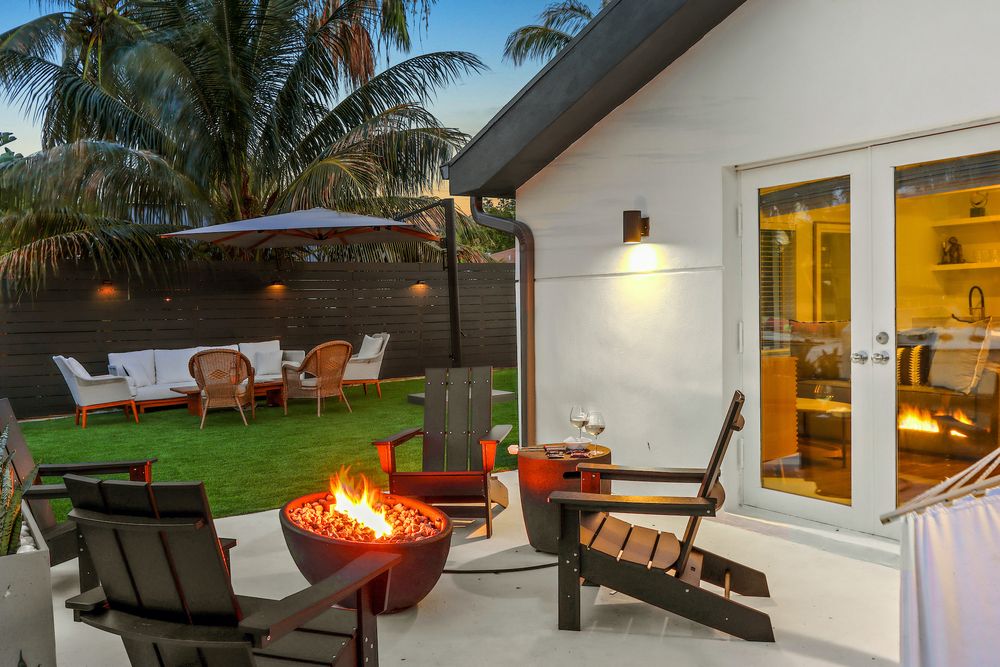 Cozy backyard patio with fire pit, lounge chairs, and palm trees illuminated at dusk with warm interior lights glowing.