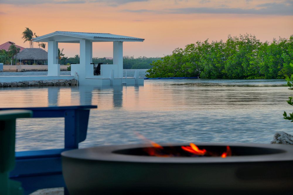 A tranquil waterfront scene at sunset with a covered boat dock and stone firepit glowing in the foreground.