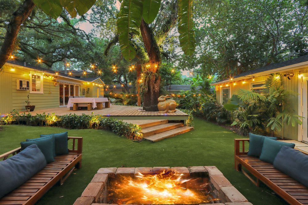Cozy backyard patio with built-in fire pit, cushioned benches, and lush landscaping illuminated by warm evening lights.