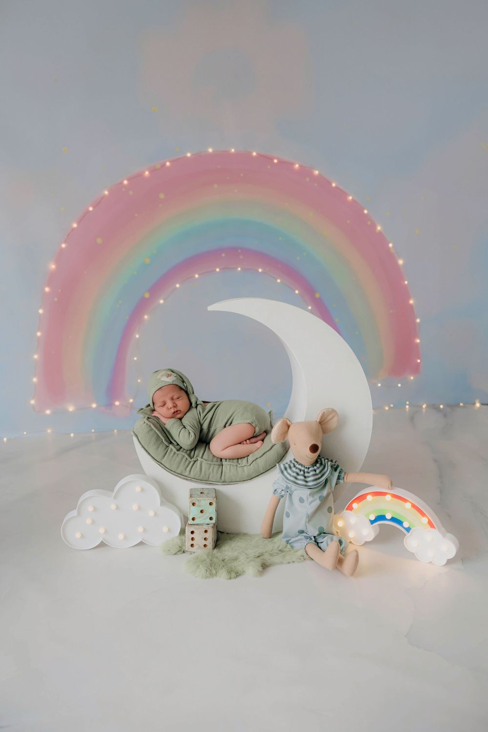 Fotografía de bebé dormido en una luna con fondo de arcoíris en su sesión en estudio.
