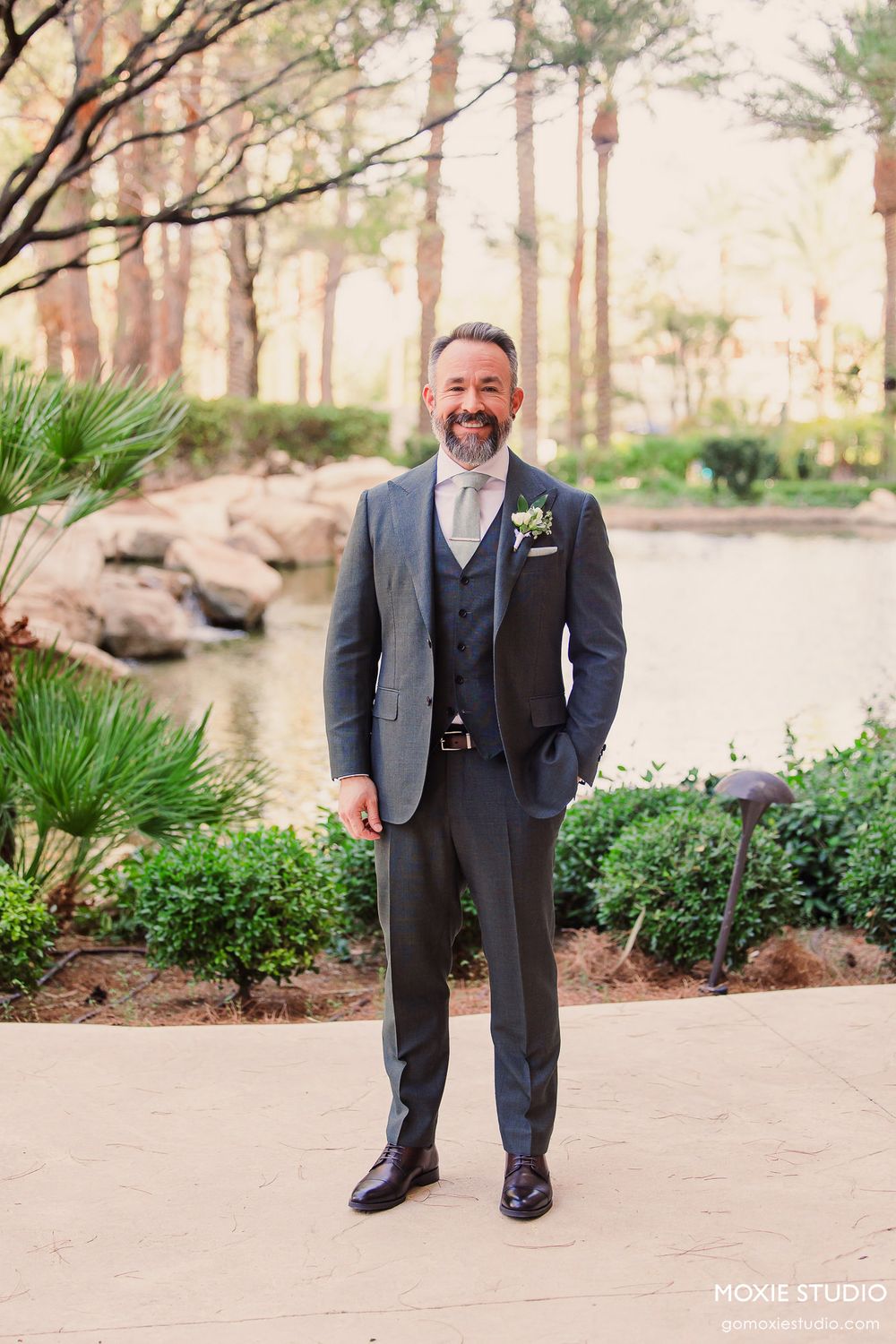 A groom in a dark grey three-piece suit with boutonniere stands on a garden path near rocks and tropical plants.