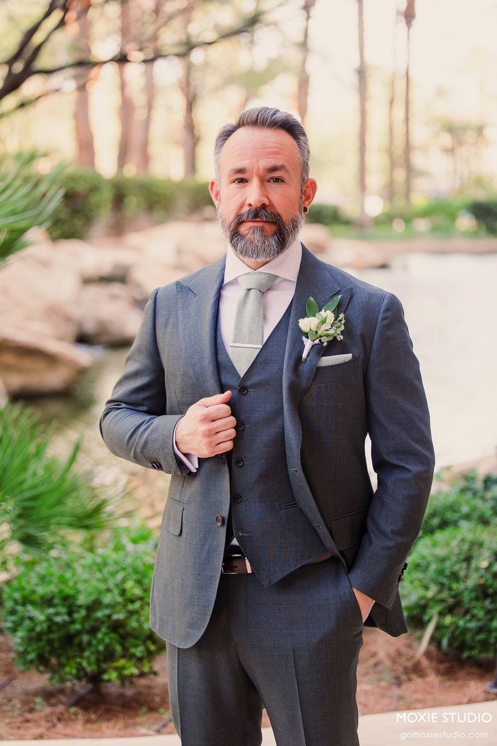 A person in a tailored grey three-piece suit with boutonniere poses outdoors among trees and greenery.