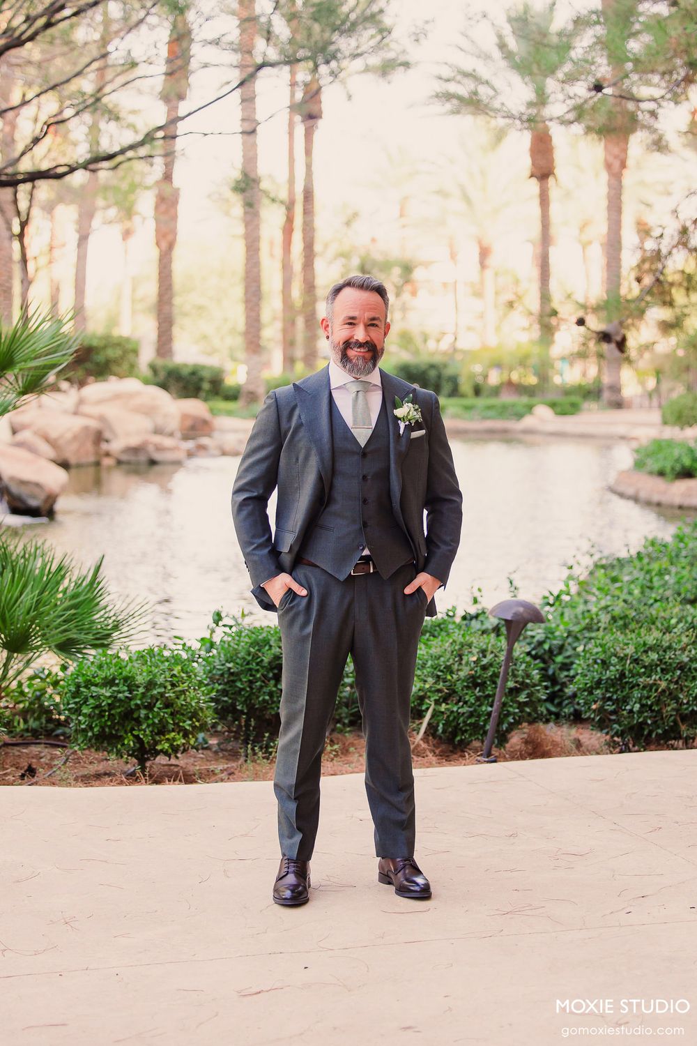 Groom in navy suit and grey vest stands confidently in outdoor garden setting with palm trees and natural landscaping.