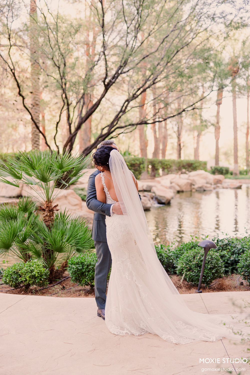 A romantic wedding moment captured from behind as a couple embraces near a serene pond with palm trees and soft lighting.