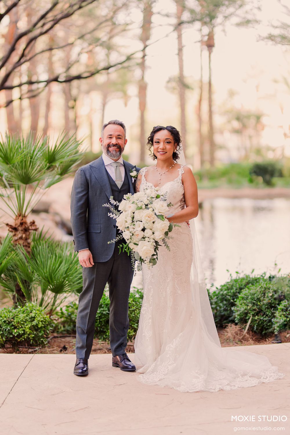 A couple poses together at their outdoor wedding ceremony with palm trees and greenery in the background.