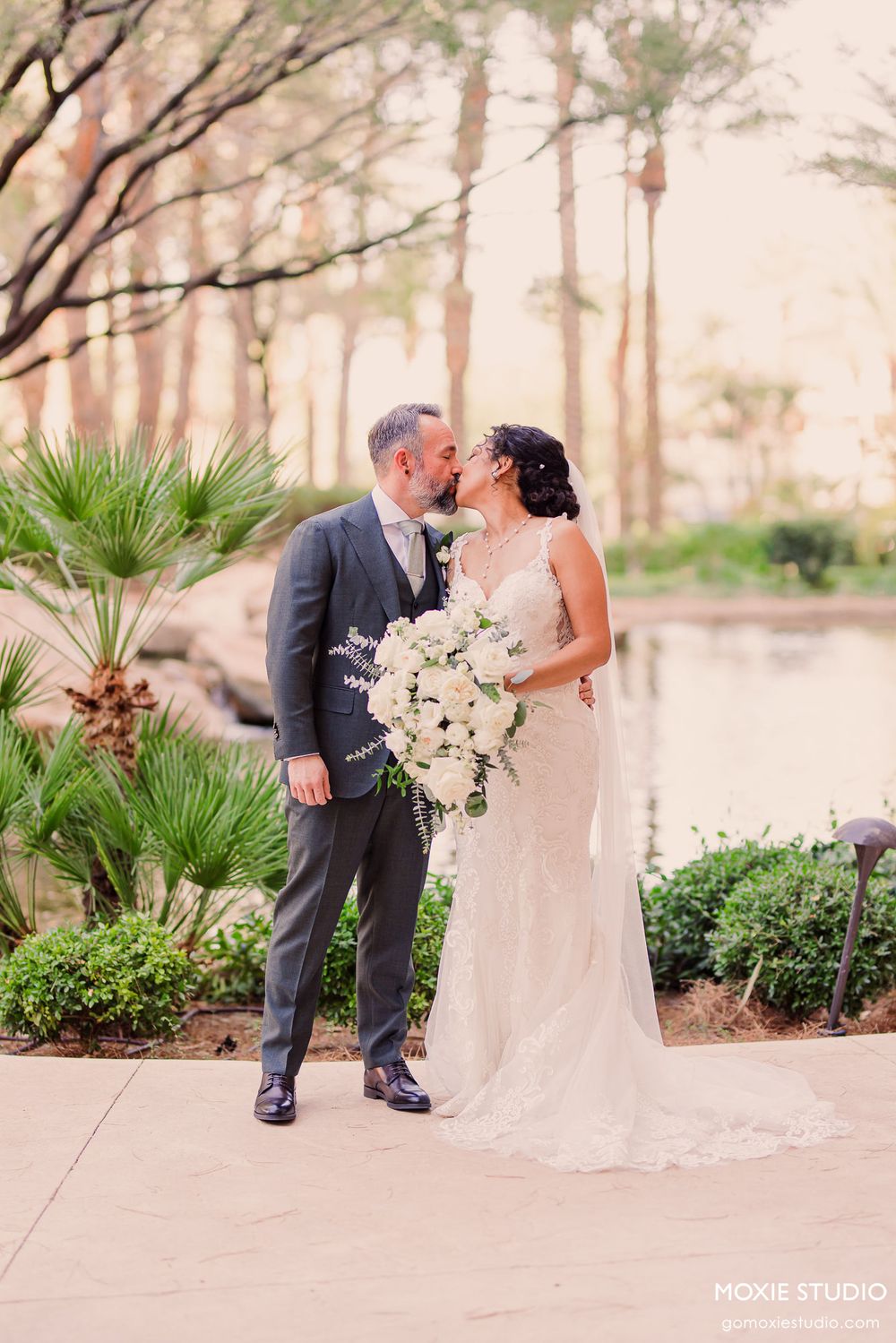 A wedding couple shares an intimate moment in a garden setting with palm trees and a beautiful floral bouquet.