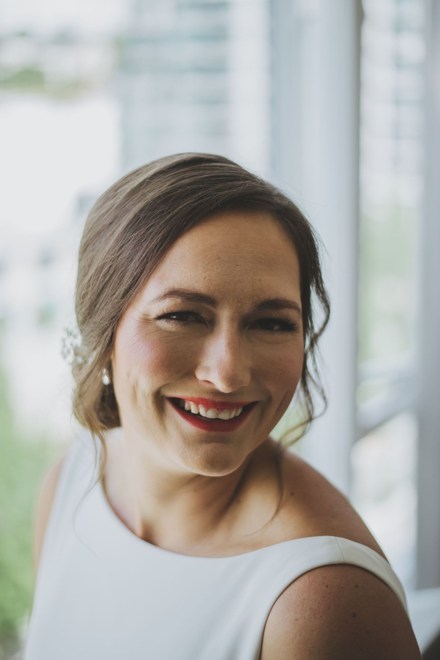 Elegant bride in white sleeveless dress smiling by bright window.