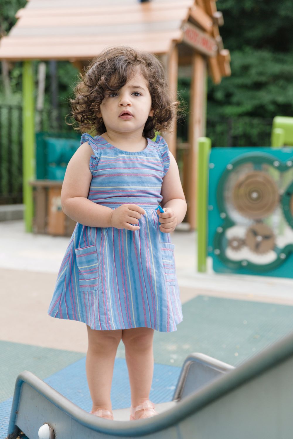 Small child wearing a striped blue dress stands on playground equipment at a park on a sunny day.