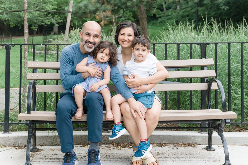 A family of four sits together on a wooden park bench surrounded by lush green foliage on a sunny day.
