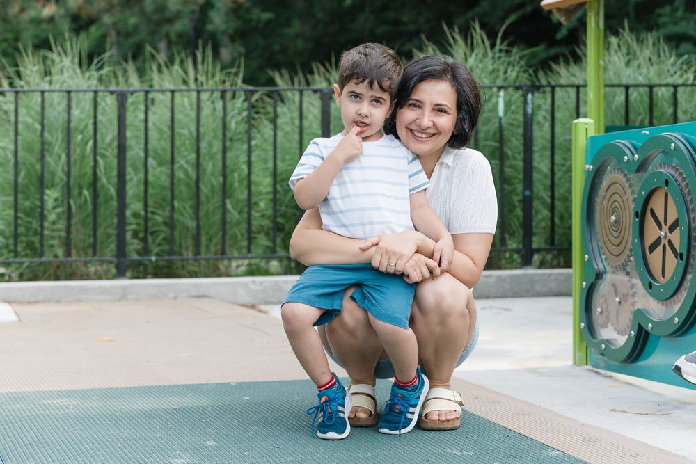 Two people sit on the ground playing and smiling together near playground equipment at a park.