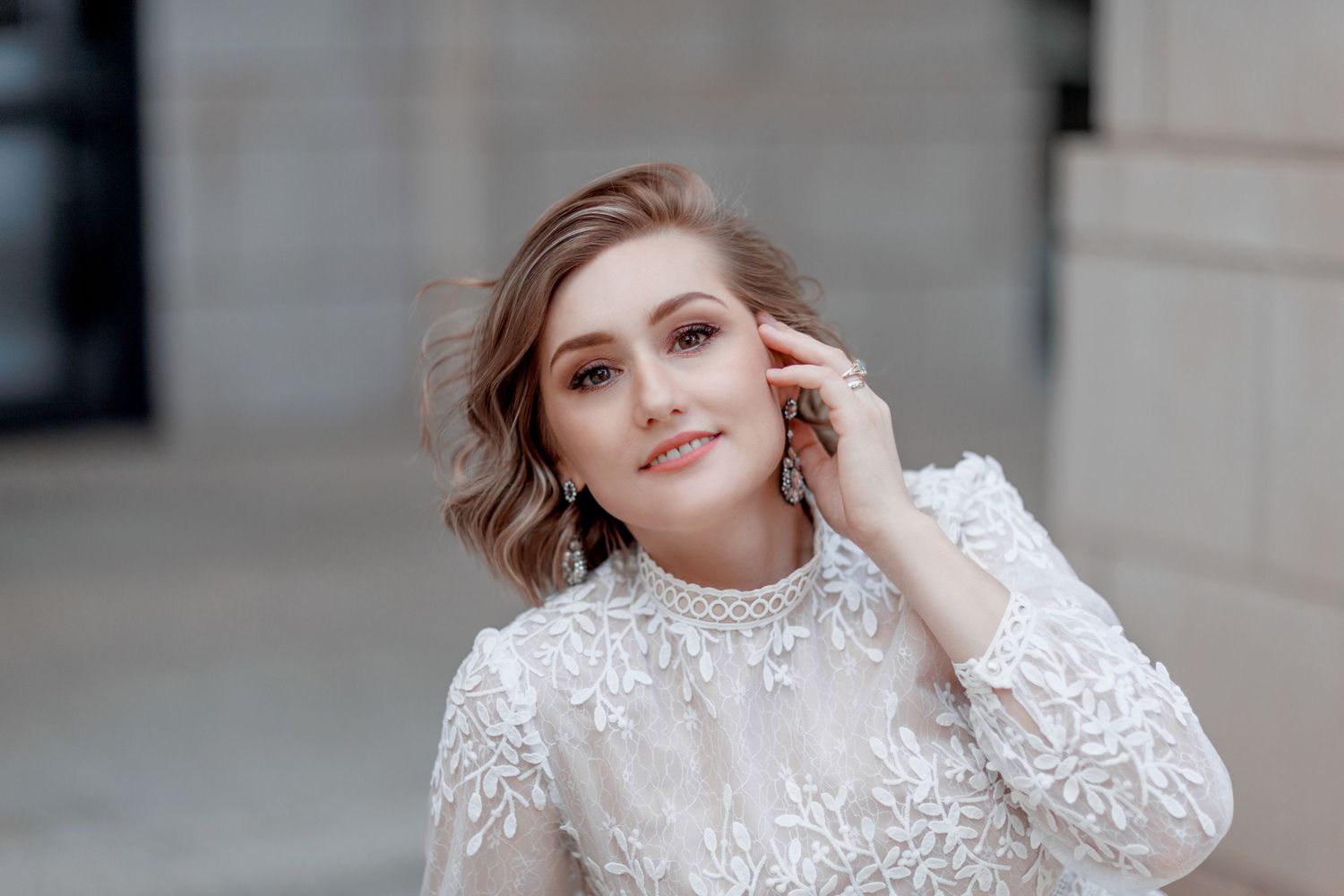A person wearing an elegant white lace dress with a high neckline poses against a neutral architectural background.