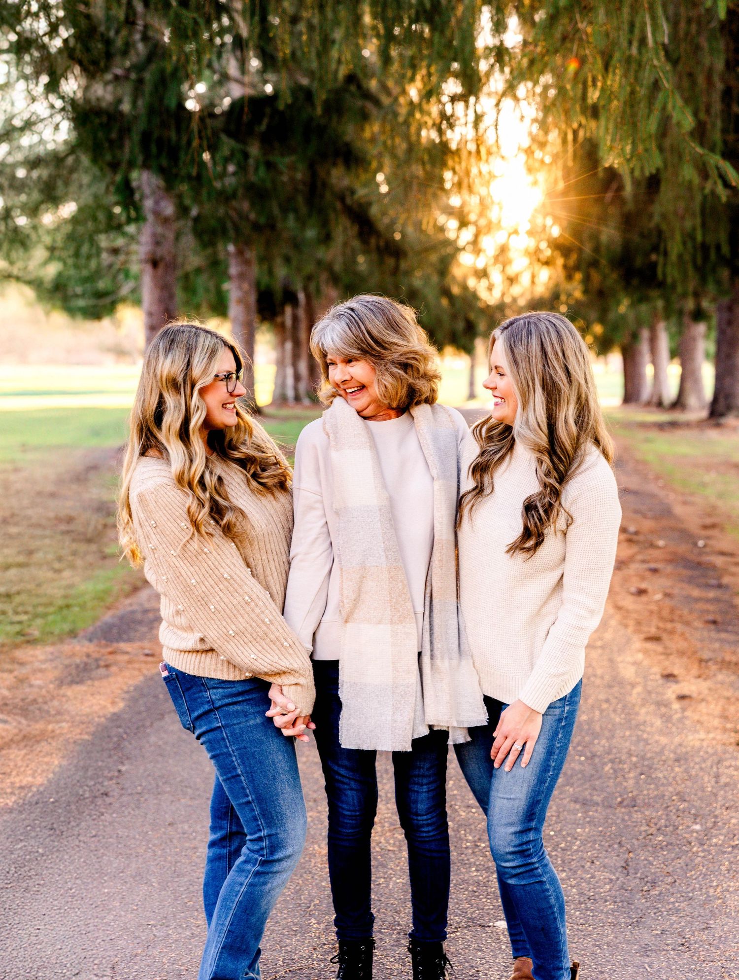 Three women in cream sweaters laugh together on tree-lined path at sunset.