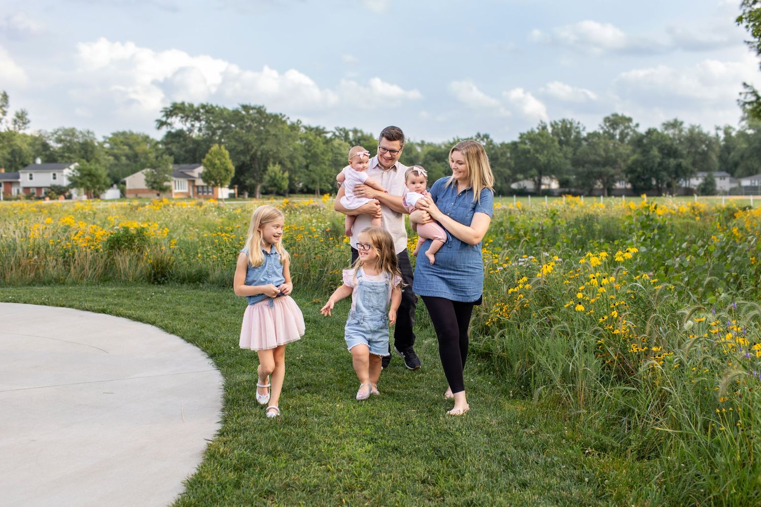 Happy family with three young daughters and baby walk along garden path surrounded by wildflowers outdoors.