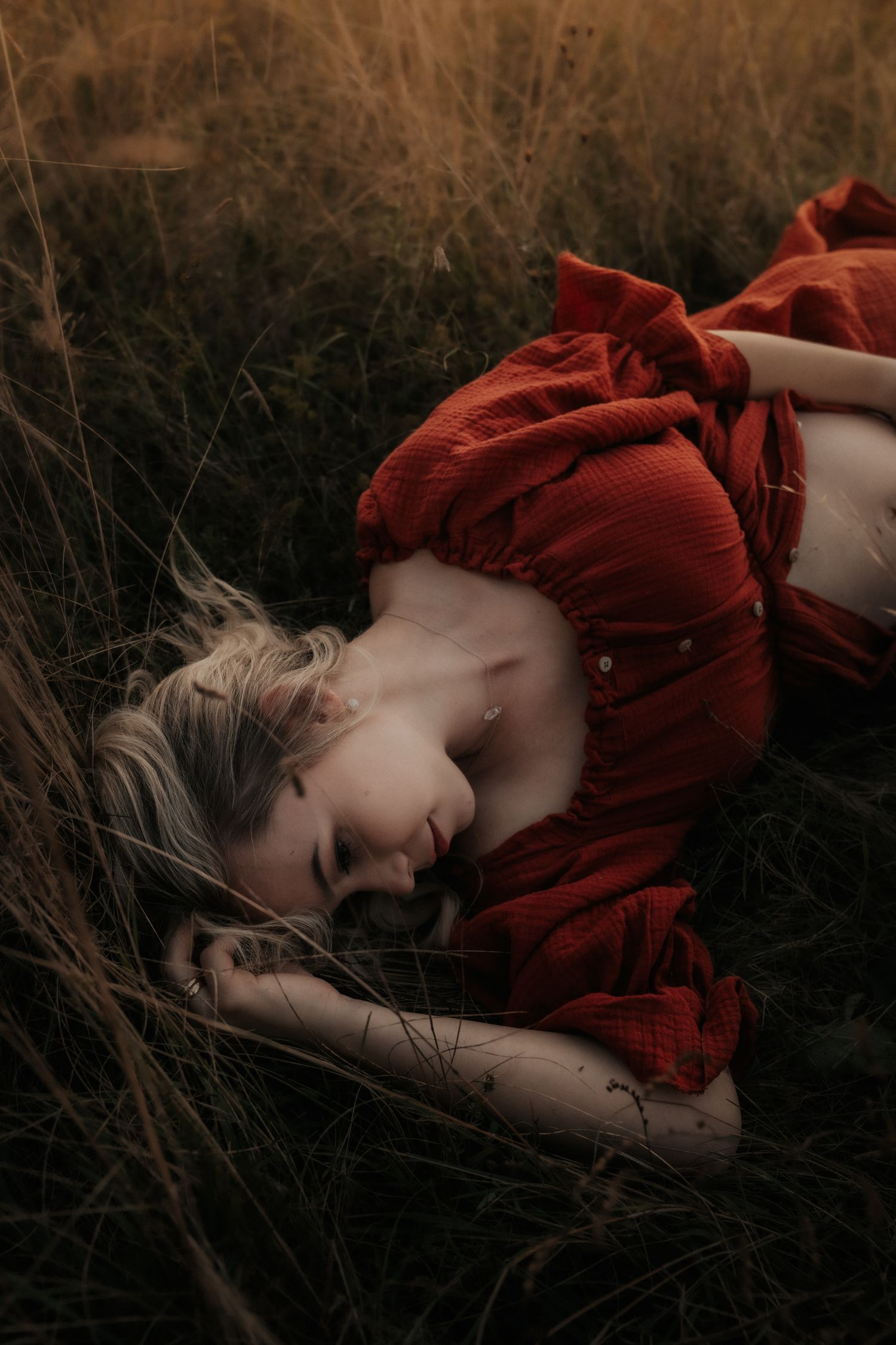 A figure in a flowing red dress lies dramatically in tall grass during golden hour.