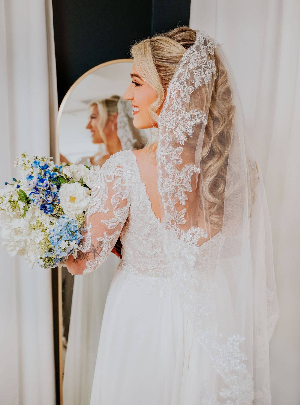 Bride in lace wedding gown with veil holds blue and white bouquet, reflected in round mirror.