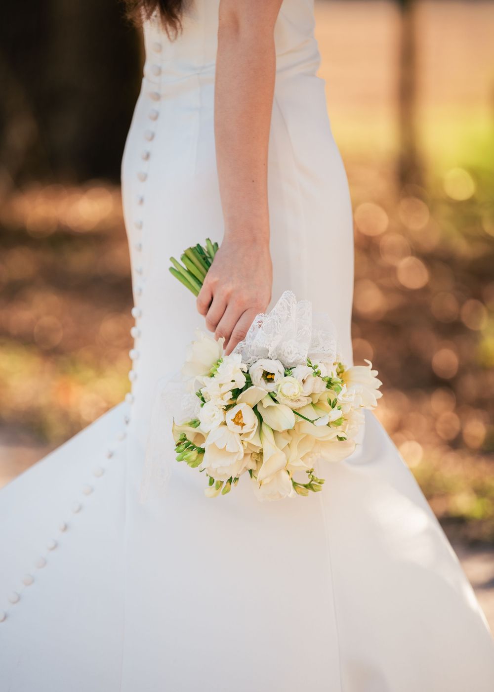 Bride in white wedding gown with button back detail holding elegant white floral bouquet outdoors.