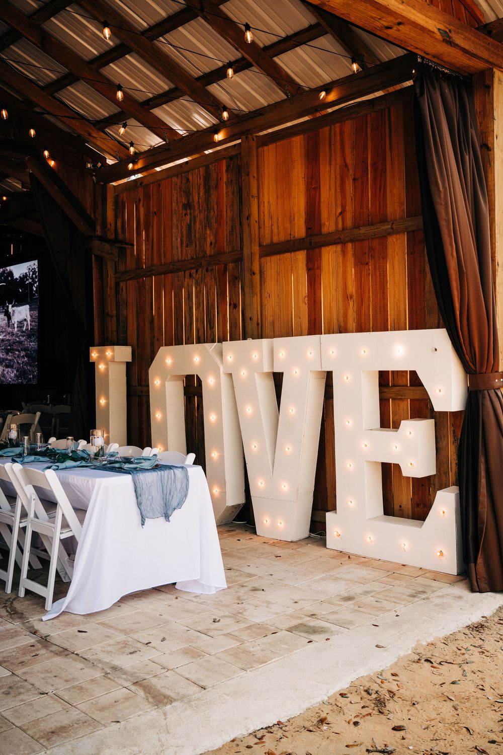 Large illuminated LOVE letters beside a decorated wedding reception table in a rustic barn venue with string lights.