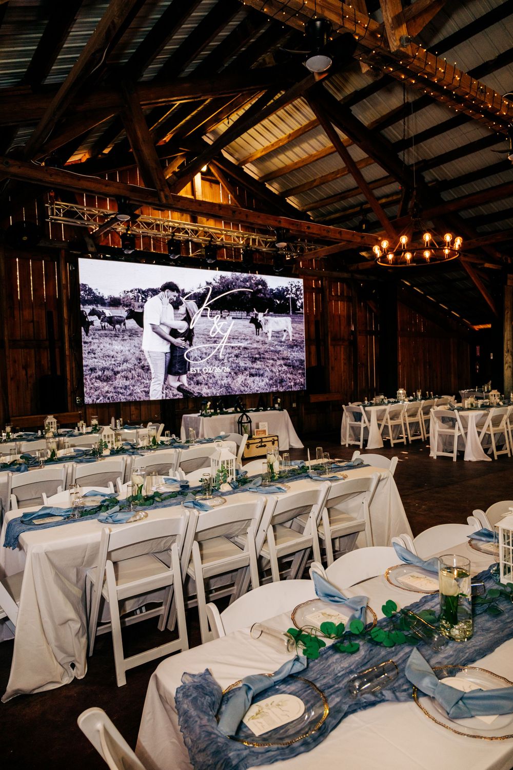 Rustic barn wedding reception with white round tables, blue napkins, and a large screen displaying a black-and-white couple photo.