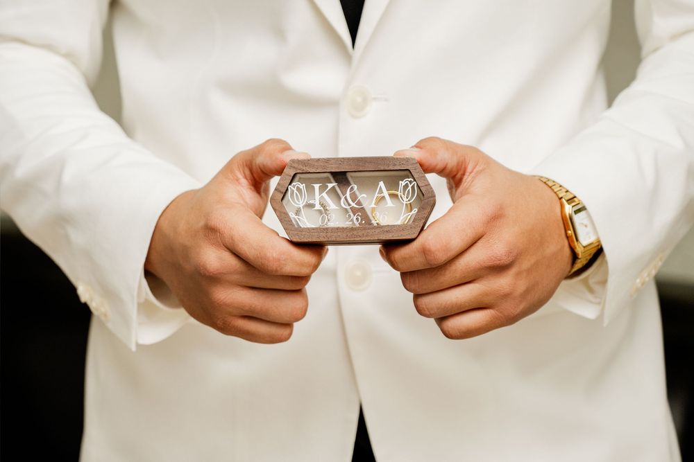 Groom in white suit holding monogrammed silver ring box with wedding bands at ceremony.