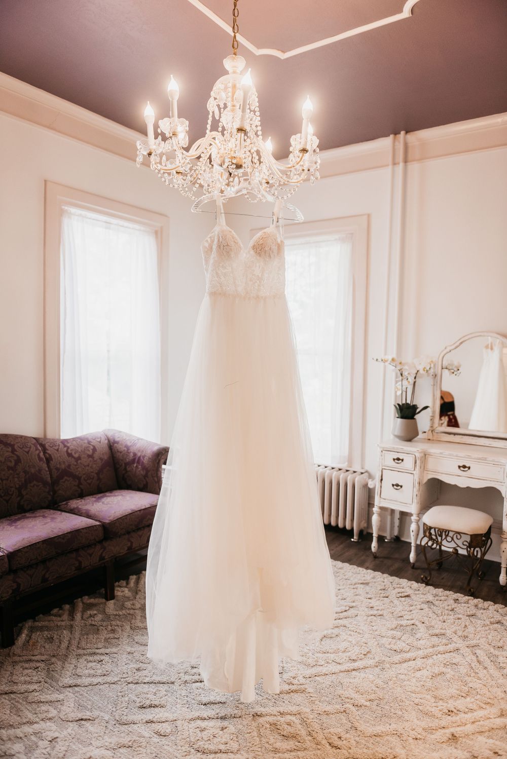 An elegant white wedding dress hangs from a crystal chandelier in a room with soft pink walls.