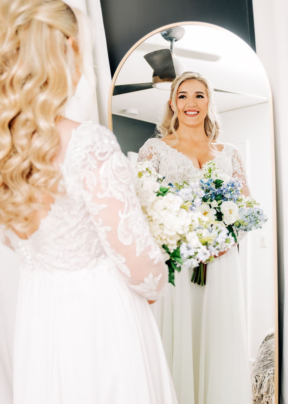Bride in lace long-sleeve wedding gown holds blue and white bouquet while admiring her reflection in an arched mirror.
