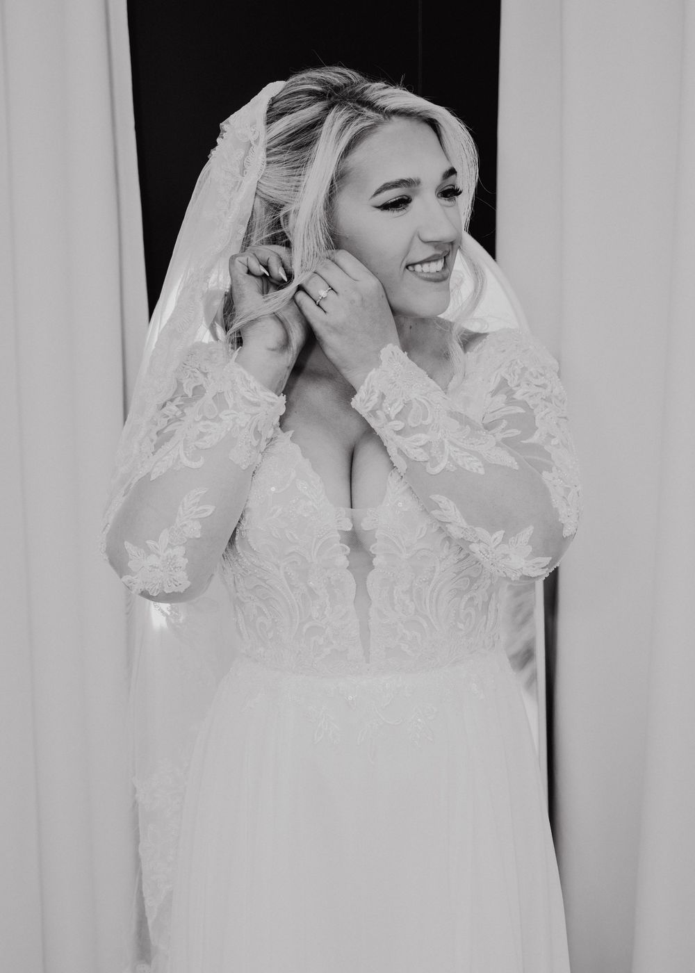 Black and white photo of a smiling bride in lace wedding dress and veil putting on earrings.