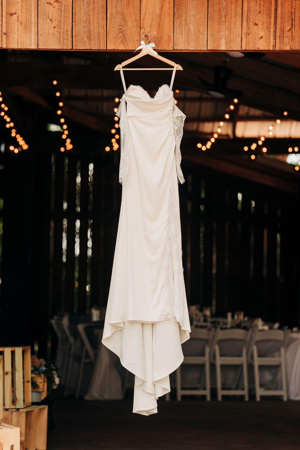 White strapless wedding gown hanging on wooden hanger in rustic barn venue with warm string lights in background.
