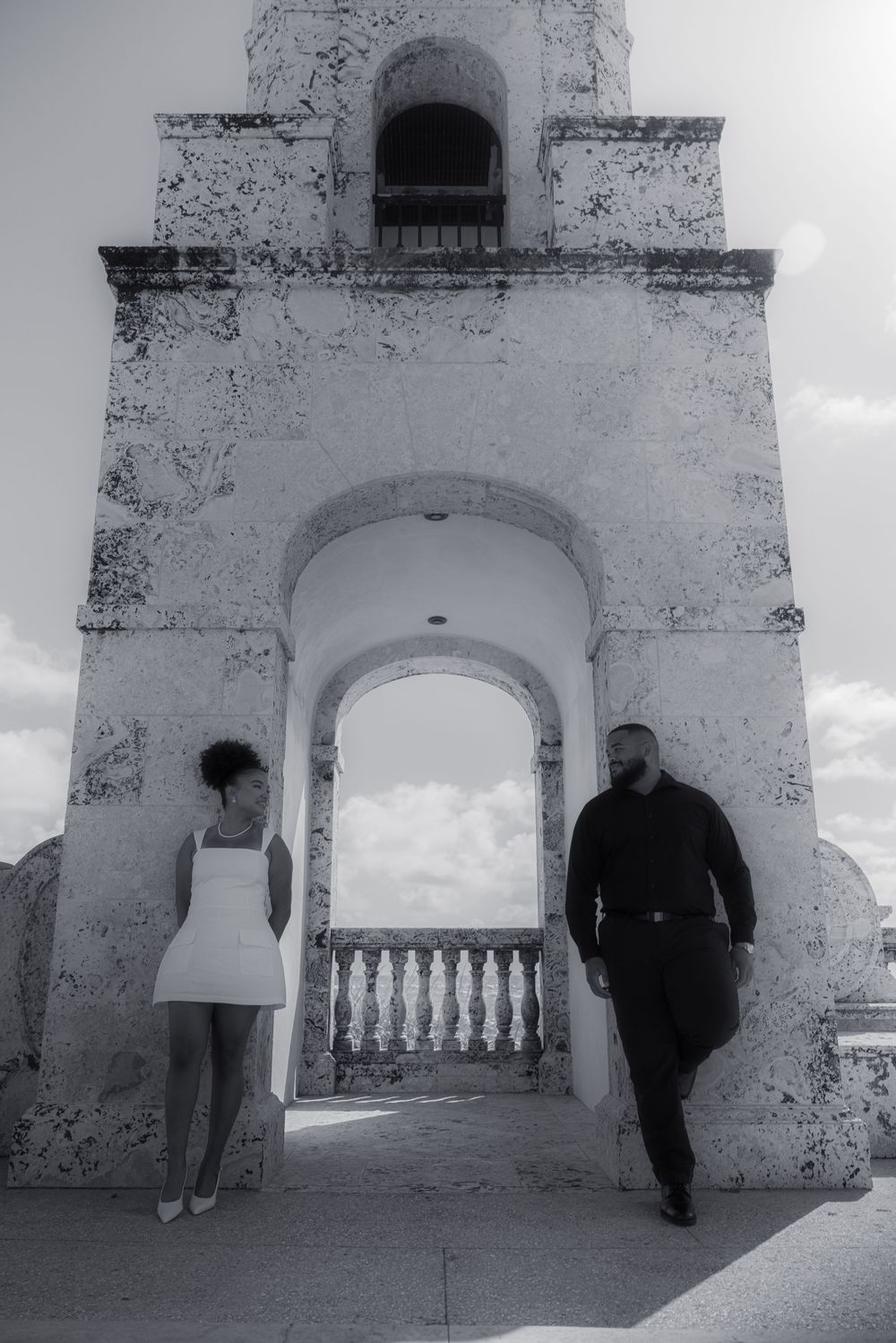 Black and white photo of a couple posing under a historic stone arch with balustrade and bell tower.