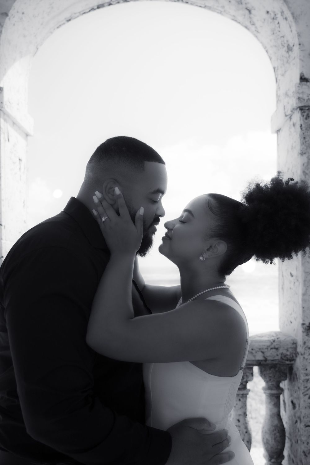 Black and white photo of a couple sharing a romantic kiss under a stone arch with ocean views.