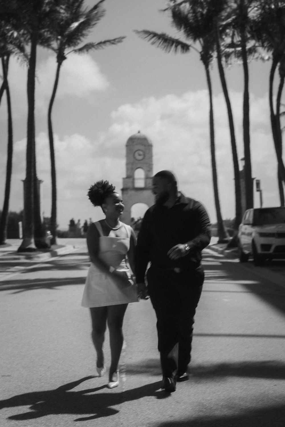 Black and white photo of a couple holding hands walking on a palm-lined street with a clock tower backdrop.