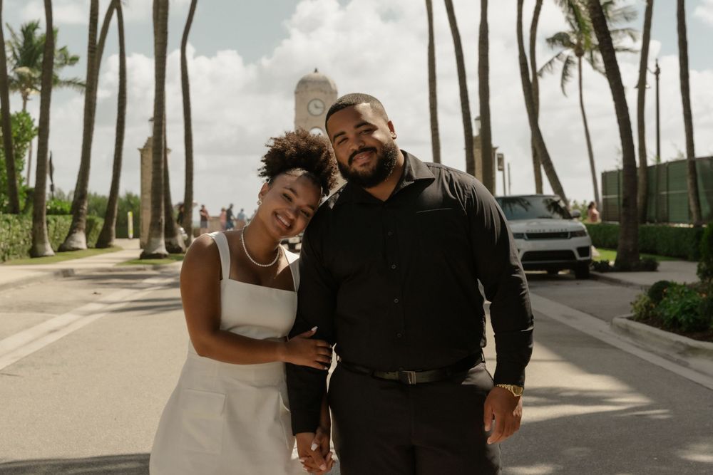 A smiling couple poses outdoors near palm trees and a white car, dressed in elegant black and white attire.