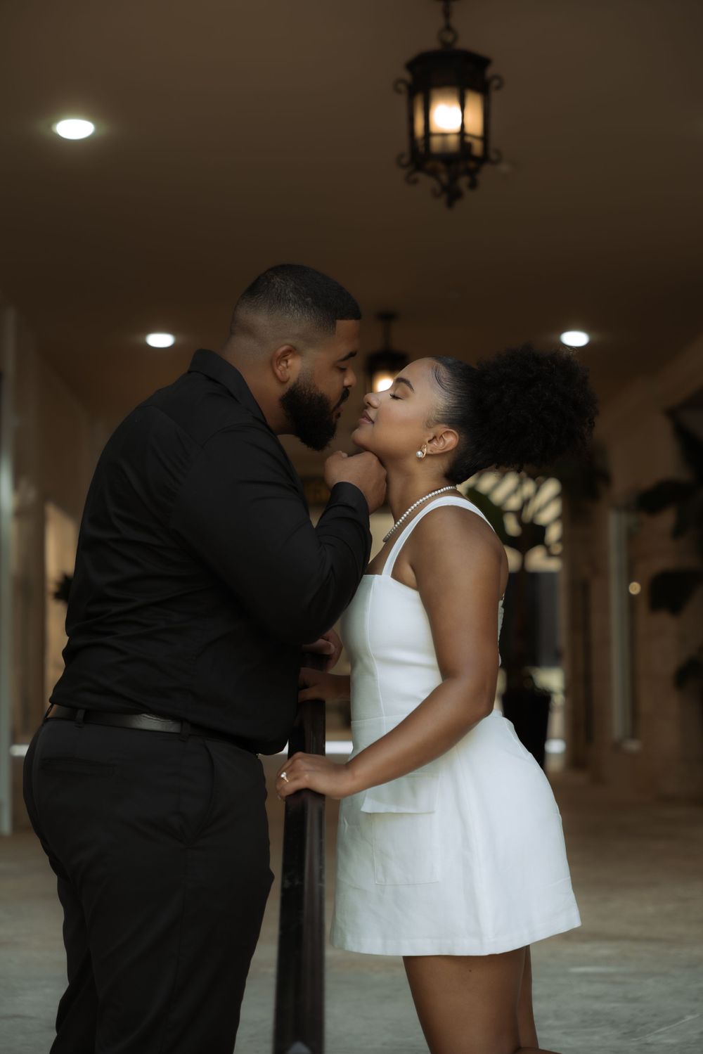 A couple sharing a romantic moment, man in black shirt and woman in white dress, under elegant chandelier lighting.
